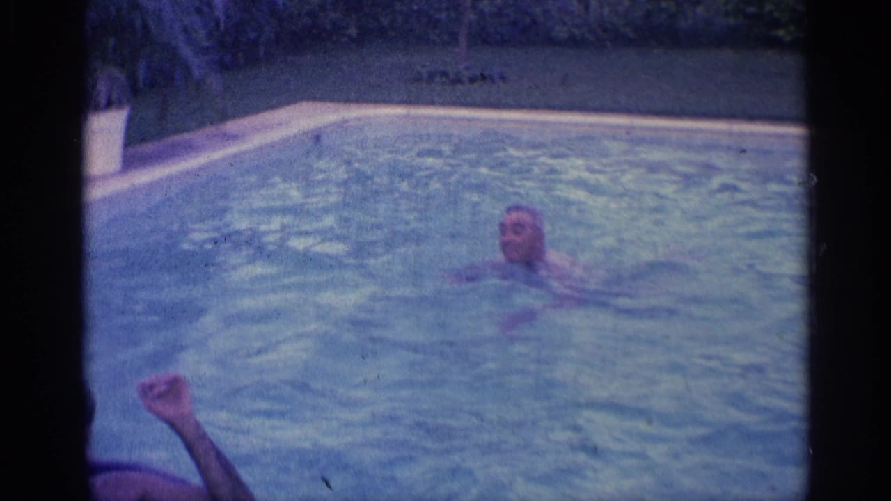Man swims in a backyard pool while another arm appears near the foreground.