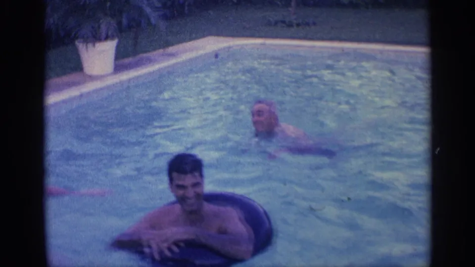 In the backyard pool, one man rests on a black inner tube in the foreground while another swimmer moves through the water behind him.
