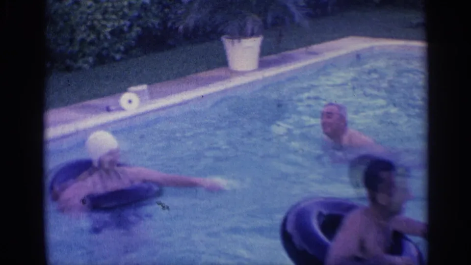 The frame shows the woman in a white swim cap floating on a tube and a man on another tube in the 1959 Indiana backyard pool scene.