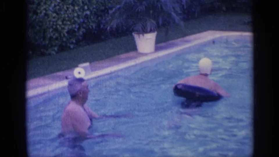 1959 Indiana home-movie frame with two adults in a backyard pool, one wearing a swim cap and floating in a dark ring.