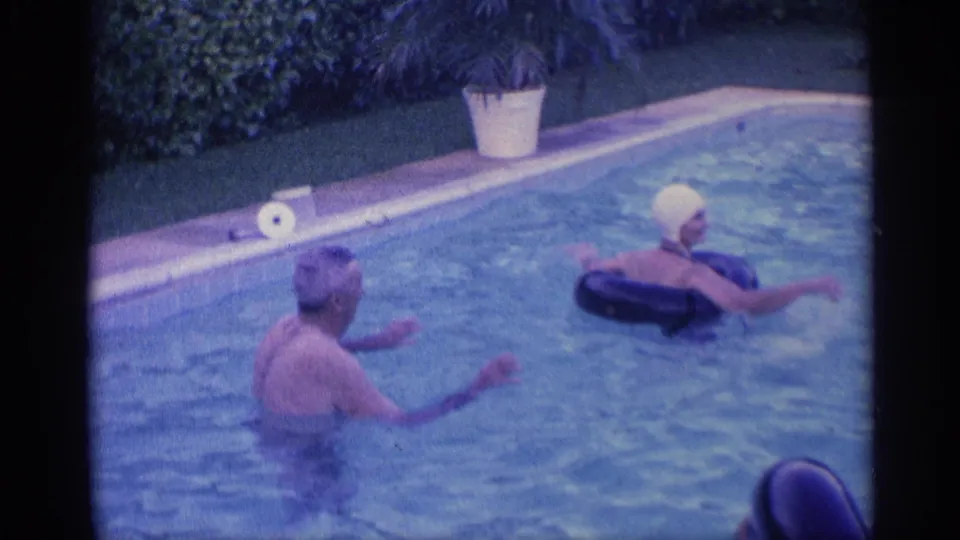 1959 Indiana backyard pool frame showing two adults in the water, one in a swim cap, with a roll on the pool edge behind them.