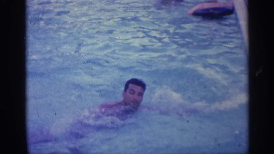 A man swims across the blue-tinted backyard pool, facing near the camera in this Indiana home-movie frame from 1959.