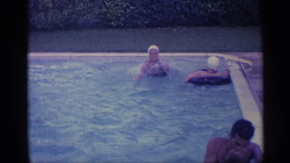 Two capped swimmers remain near the far pool edge as another man appears at the lower right in this blue-tinted Indiana 8mm frame.