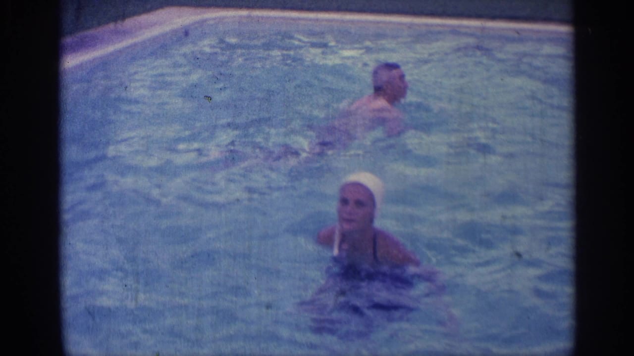 Two adults swim in a backyard pool, with one wearing a white swim cap.