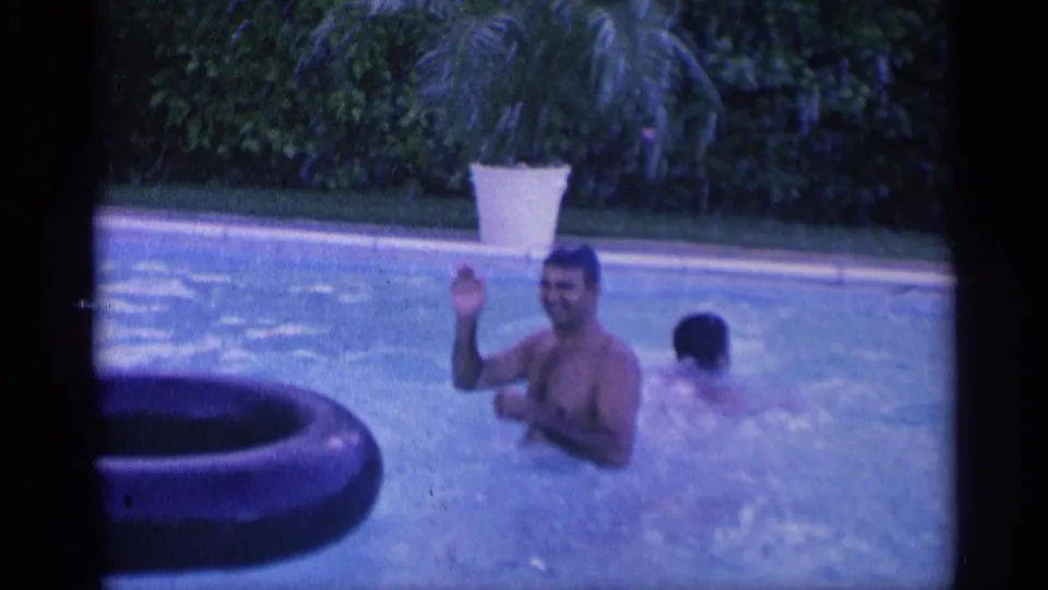 1959 Indiana 8mm frame of a smiling adult man waving from a backyard pool as another swimmer moves behind him.