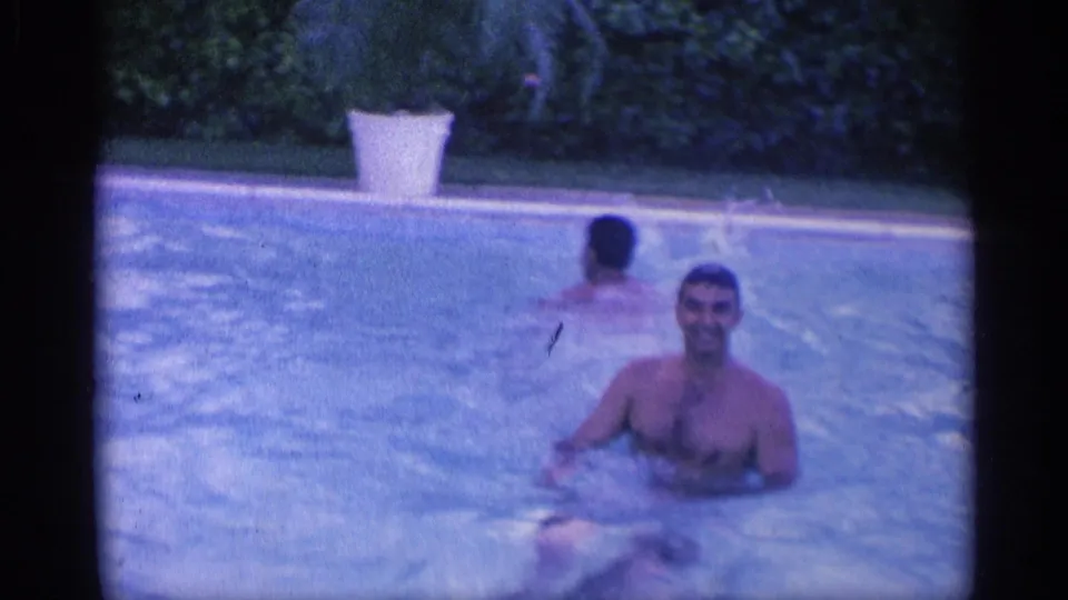 1959 Indiana home-movie frame of a smiling man in a backyard pool with another swimmer turned away near the far side.