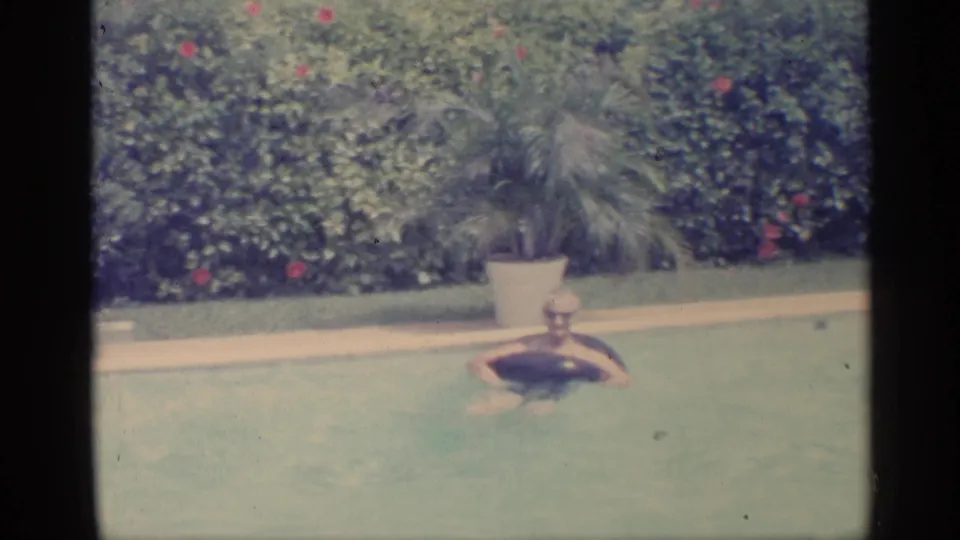 Indiana 1959 home-movie frame showing a dark inflatable ring in a backyard pool near a potted palm and flowering hedge.