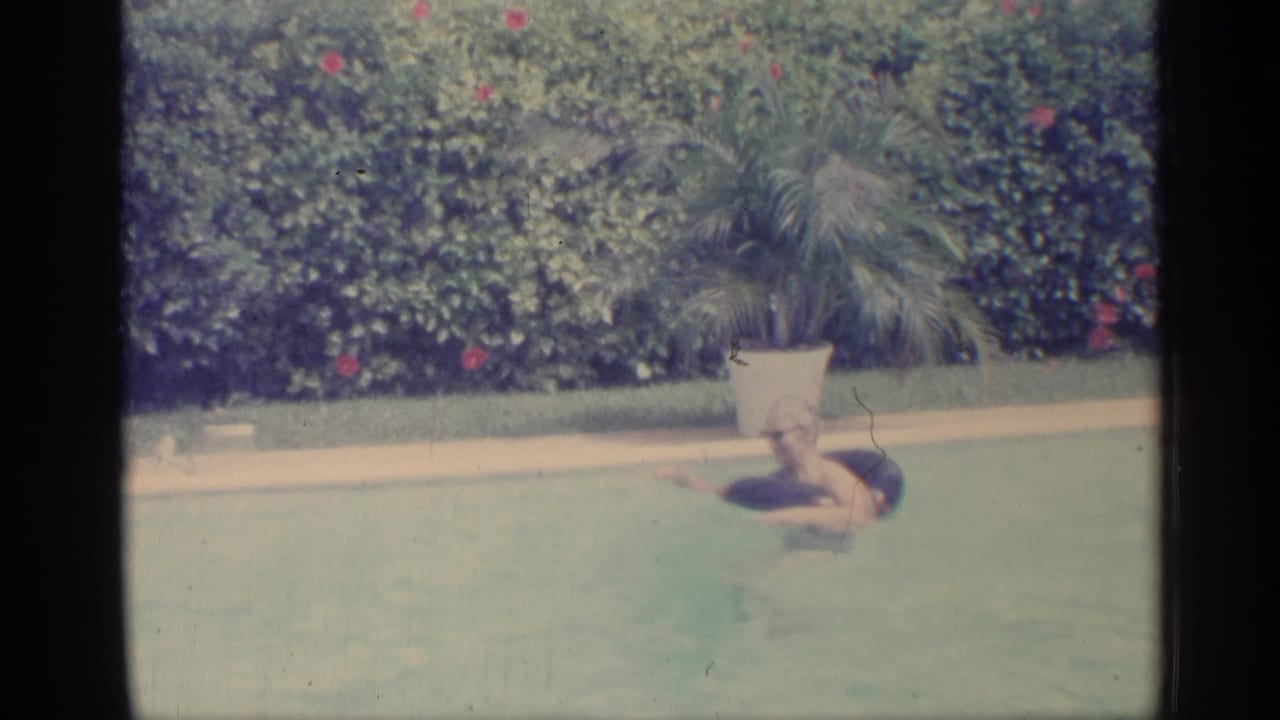 1959 Indiana 8mm frame showing a person swimming in a backyard pool beside a potted palm and flowering hedge.