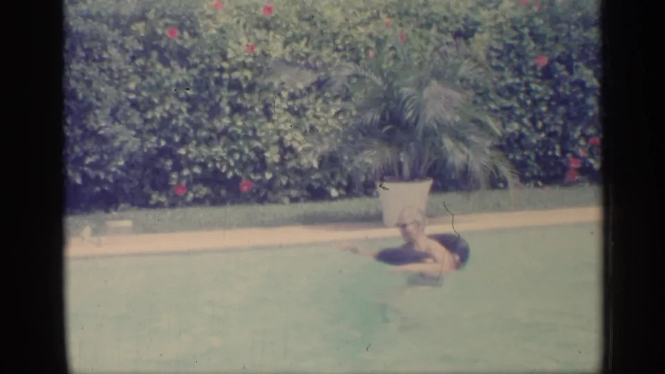 1959 Indiana 8mm frame showing a person swimming in a backyard pool beside a potted palm and flowering hedge.