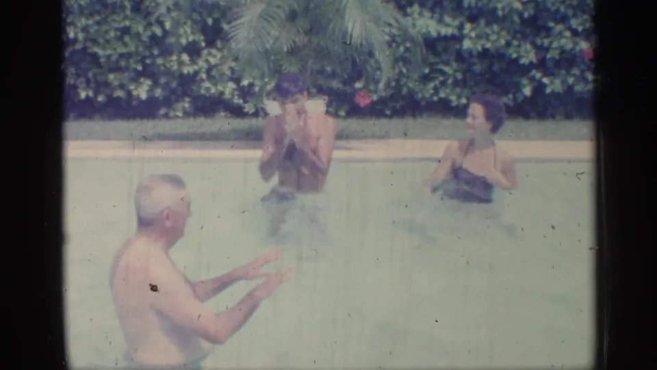 1959 Indiana 8mm frame of family members standing in a backyard pool, gesturing and talking near a potted palm and hedge.