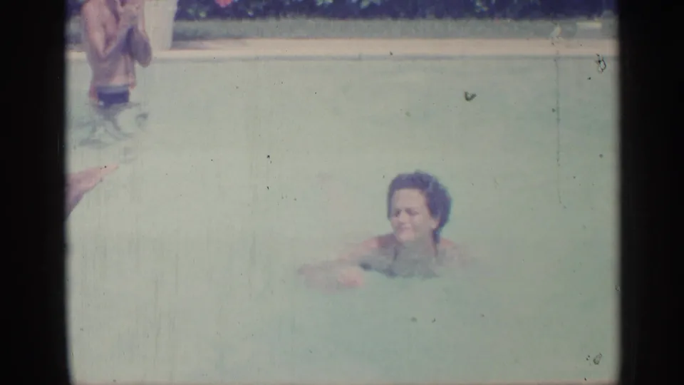 1959 Indiana home-movie frame with a woman swimming across a pale outdoor pool as another swimmer is partly cropped above.