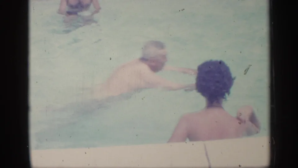1959 Indiana home-movie frame showing an older man swimming near a woman at the pool edge, with pale water filling the scene.