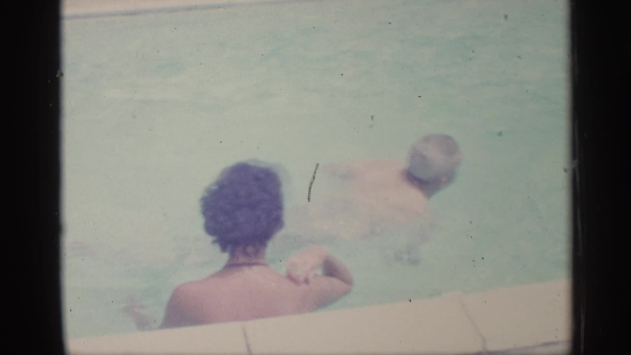Woman seated at pool edge while an adult swims nearby in pale blue water.