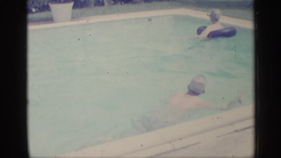 1959 Indiana home-movie frame showing an adult in a backyard pool with a child floating on a dark inner tube nearby.