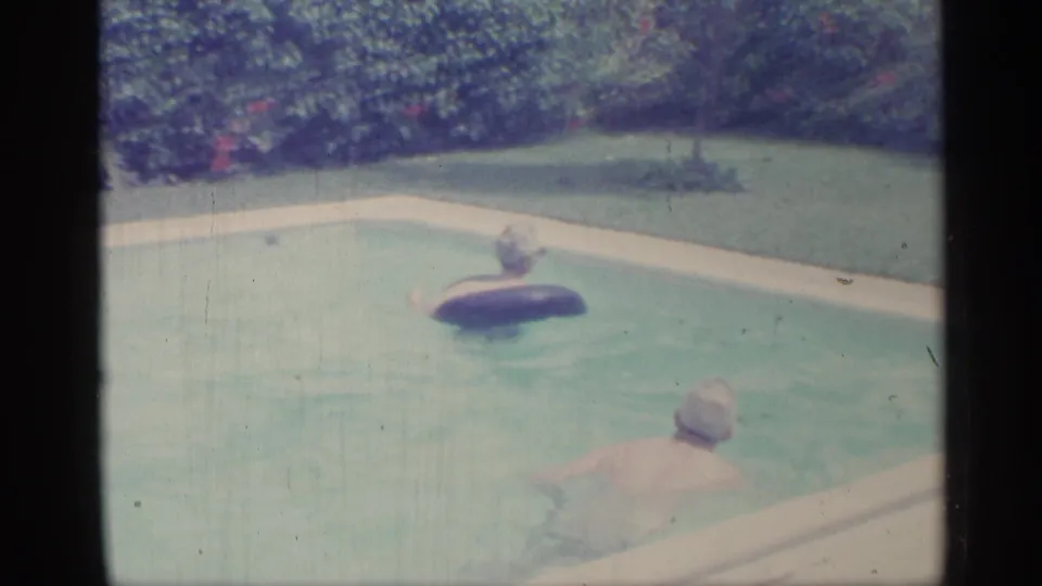 1959 Indiana home-movie frame showing a child on an inner tube and an adult swimmer in a hedge-lined backyard pool.