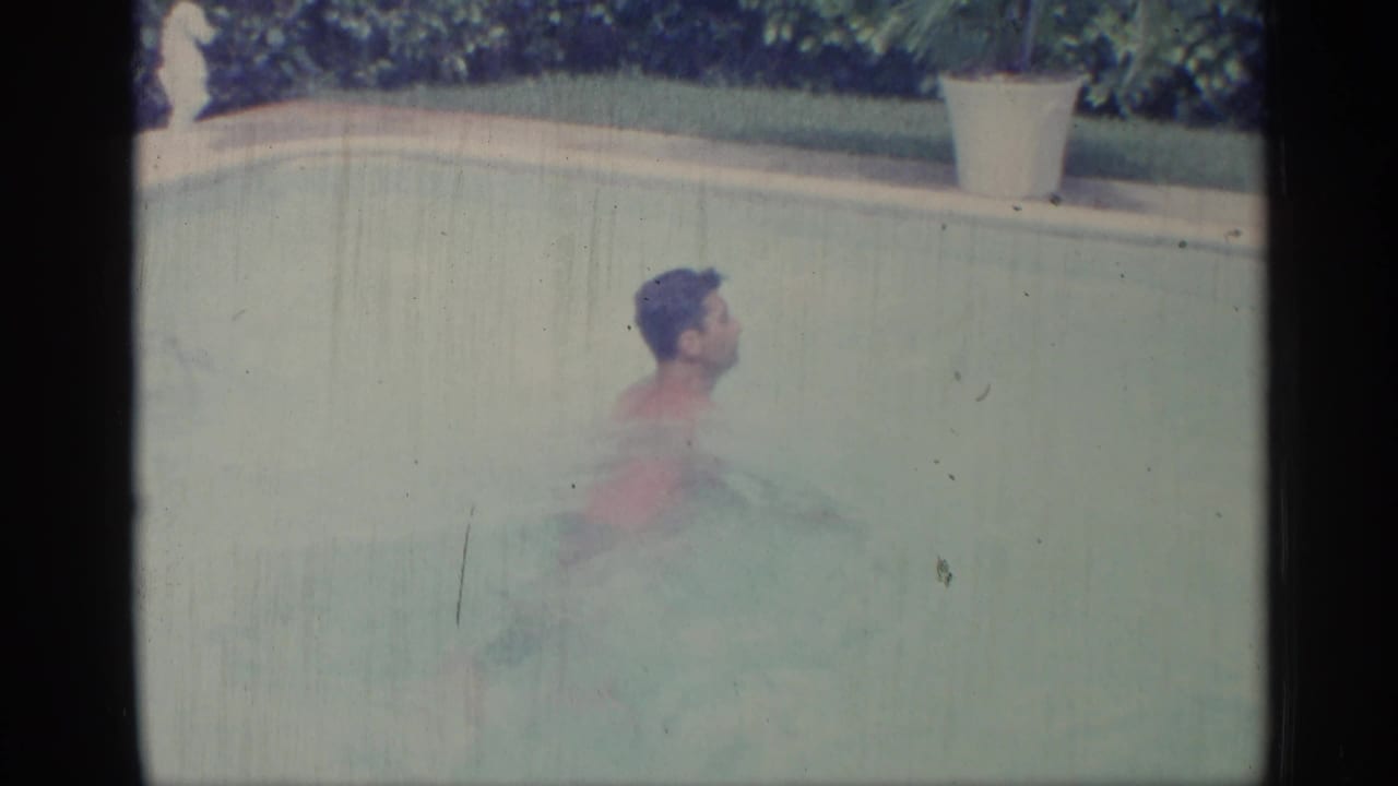 Young man swimming in a backyard pool beside pale coping and hedge foliage.