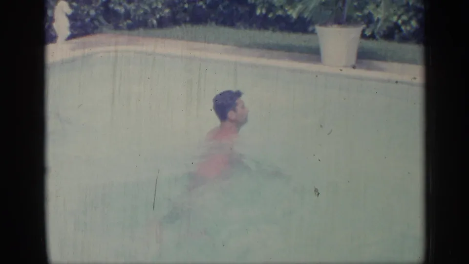 1959 Indiana 8mm frame showing a young man chest-deep in a backyard pool, with hedge greenery and a white potted plant along the edge.