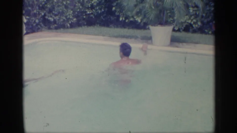 1959 Indiana home-movie frame showing the swimmer turned away in the pool, with a pale pool edge, hedge, and large white planter visible.