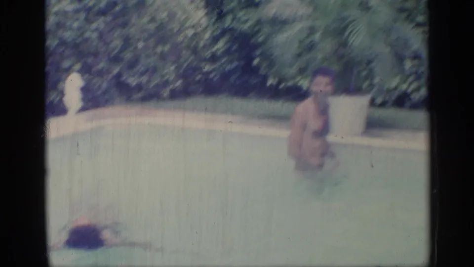 1959 Indiana 8mm frame showing a young man facing the camera in pool water, with dense greenery and a white planter behind the pool edge.