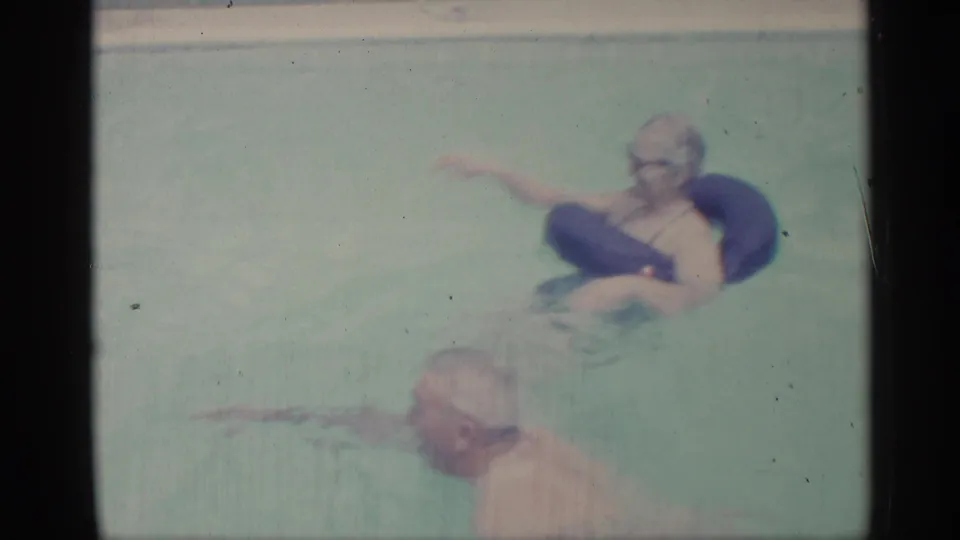 1959 Indiana home-movie frame showing two people in a backyard pool, including one reclining on a dark inflatable float and another nearer the camera.