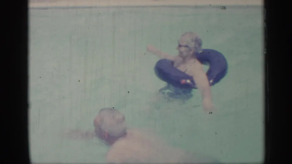 1959 Indiana home-movie frame showing a woman on a blue inner tube as an adult man swims nearby in pale pool water.