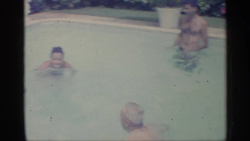 Home-movie frame from 1959 Indiana showing multiple adults in a swimming pool, including a foreground swimmer and a man farther back.
