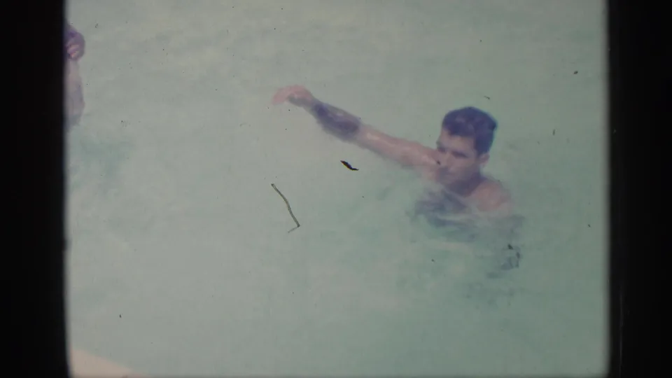 1959 Indiana home-movie frame of an adult man swimming through pale pool water, with his arm lifted mid-stroke.