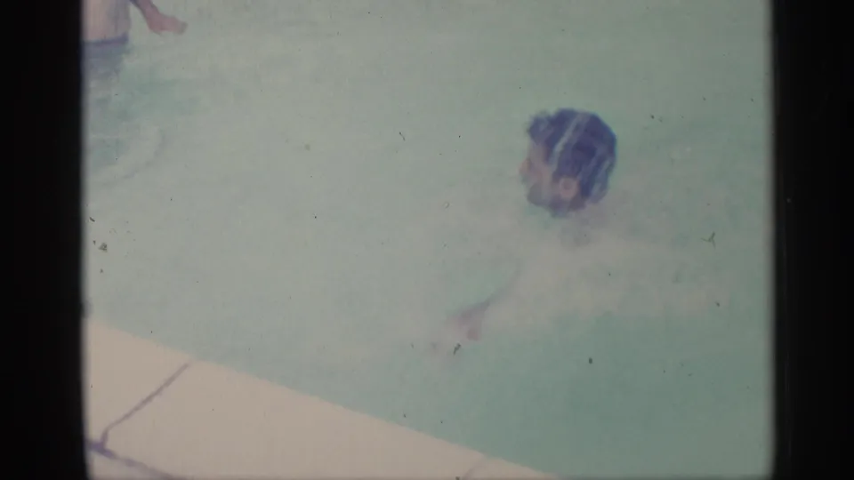 1959 Indiana home-movie frame showing a young man partly submerged in a backyard swimming pool near the pale pool coping.
