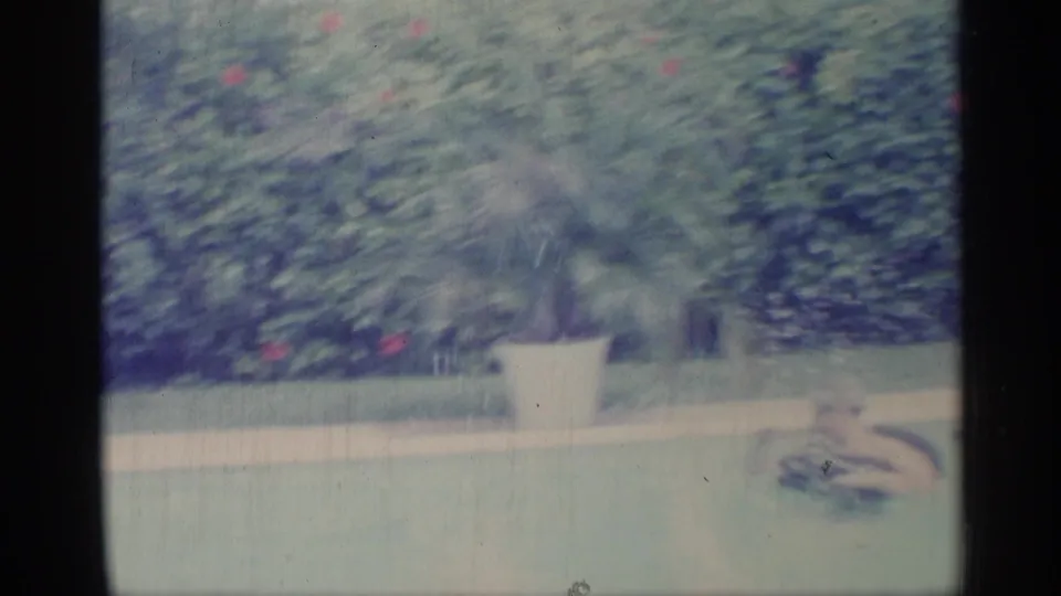 Backyard pool frame showing clipped greenery, a white planter, and a swimmer low in the water in 1959 Indiana footage.