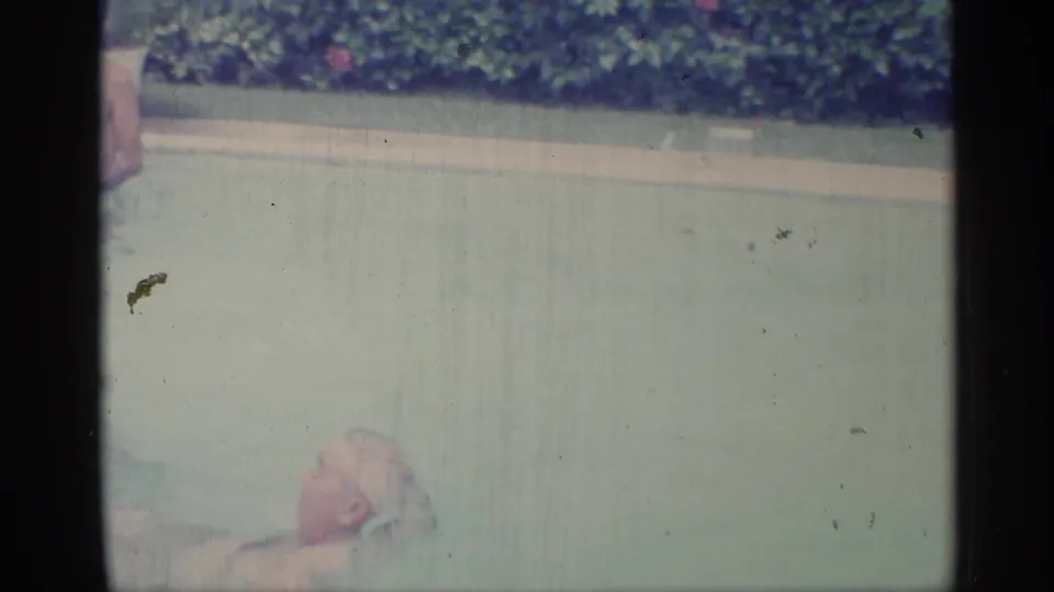 A swimmer is partly submerged near the foreground of a backyard pool, with hedges and pale coping visible in 1959 Indiana home-movie footage.