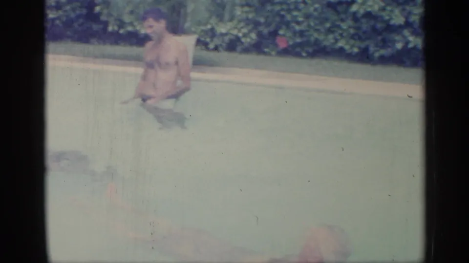 1959 Indiana 8mm frame showing an adult man waist-deep in a backyard pool with dense greenery behind him.