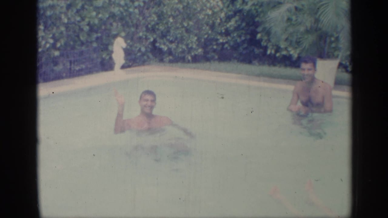 Two adult men in a swimming pool, one waving toward the camera.