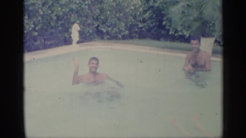 1959 Indiana home-movie frame with two adult men in a backyard pool, one smiling and waving toward the camera.