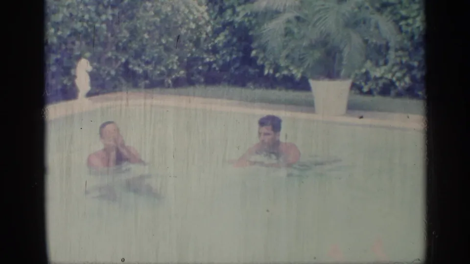 1959 Indiana 8mm frame showing two adult men swimming near a hedge-lined backyard pool edge and potted plant.