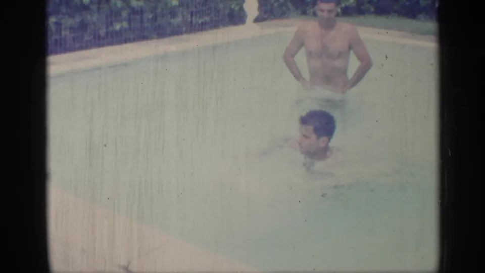 1959 Indiana home-movie frame with one adult man standing in the pool and another swimming in the foreground.