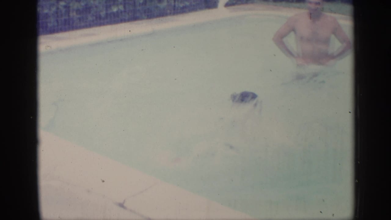 Two adult men in a swimming pool, one partly submerged and one standing near the tiled edge.