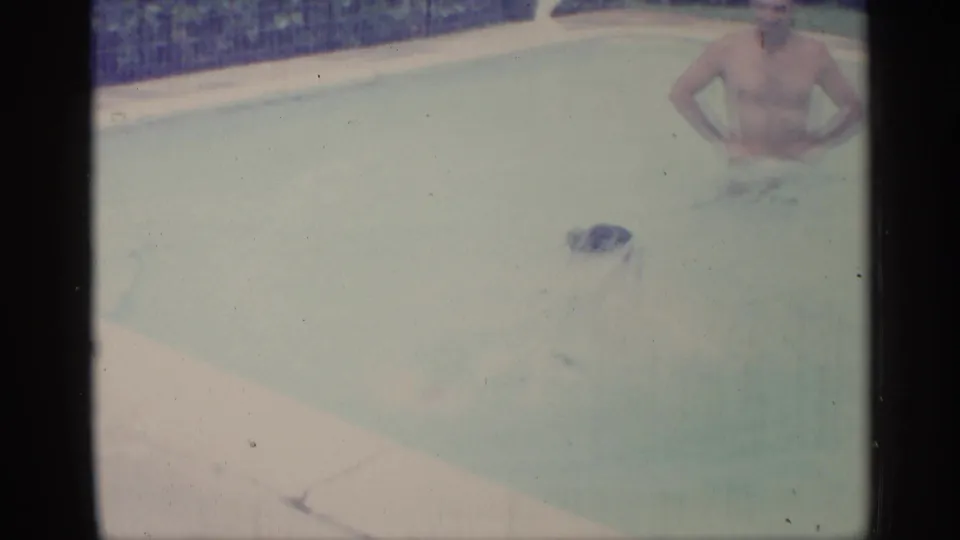1959 Indiana 8mm frame showing adult men in a backyard pool, with pale coping, blue tile, and soft faded color.
