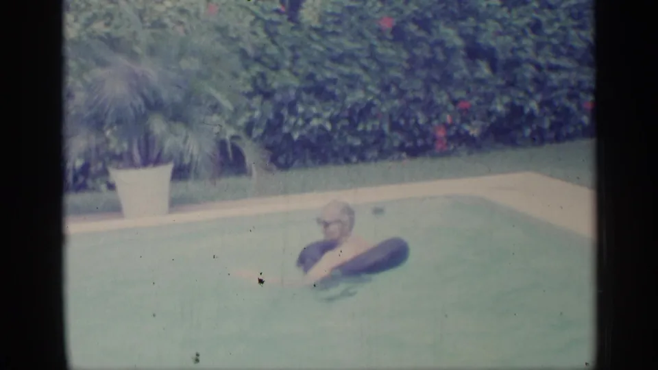 1959 Indiana home-movie frame of an adult man floating with a dark inner tube near hedges and a potted palm.