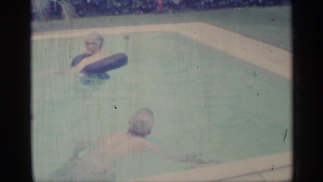 Two people in a backyard swimming pool, one floating in a dark inner tube.