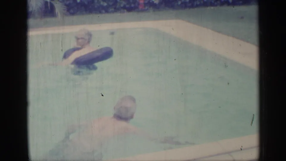 1959 Indiana home-movie frame showing one person in glasses floating on a dark inner tube while another swims nearby in a backyard pool.