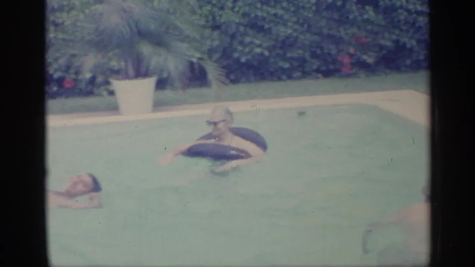 1959 Indiana home-movie frame showing two pool users, one with a dark inner tube, with hedges and a potted palm in the background.