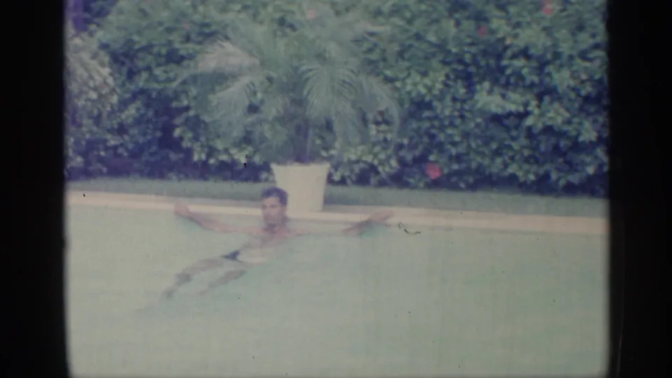 1959 Indiana home-movie frame of an adult man in a pool, arms spread along the edge, with hedges and a potted palm behind him.
