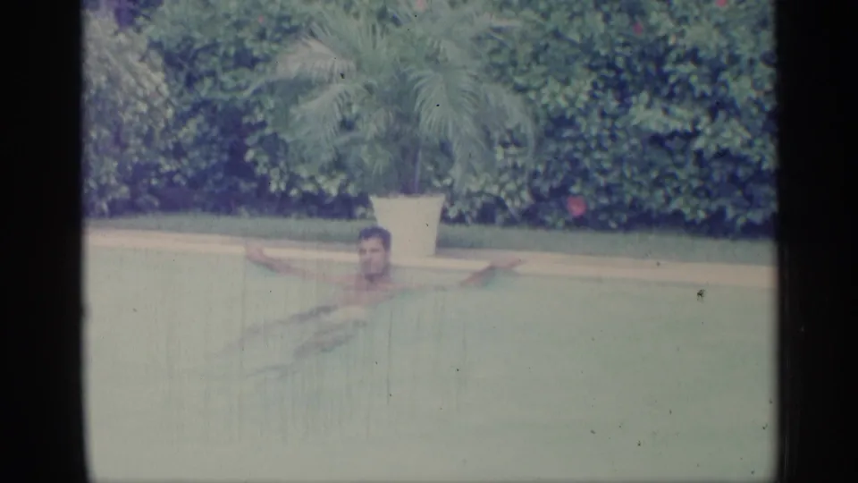 1959 Indiana 8mm frame showing an adult man swimming across blue-green pool water with hedges and a potted palm in the background.
