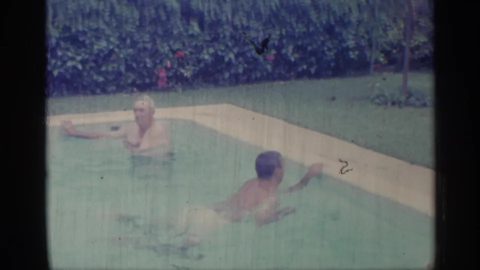 1959 Indiana home-movie frame with two adult men in a backyard-style pool, one standing or resting near the edge as another swims nearby.