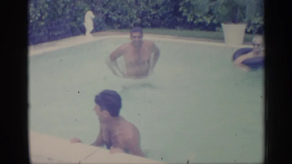 1959 Indiana 8mm frame showing adult men in a backyard pool, with one near the edge and others in the water before dense hedges.