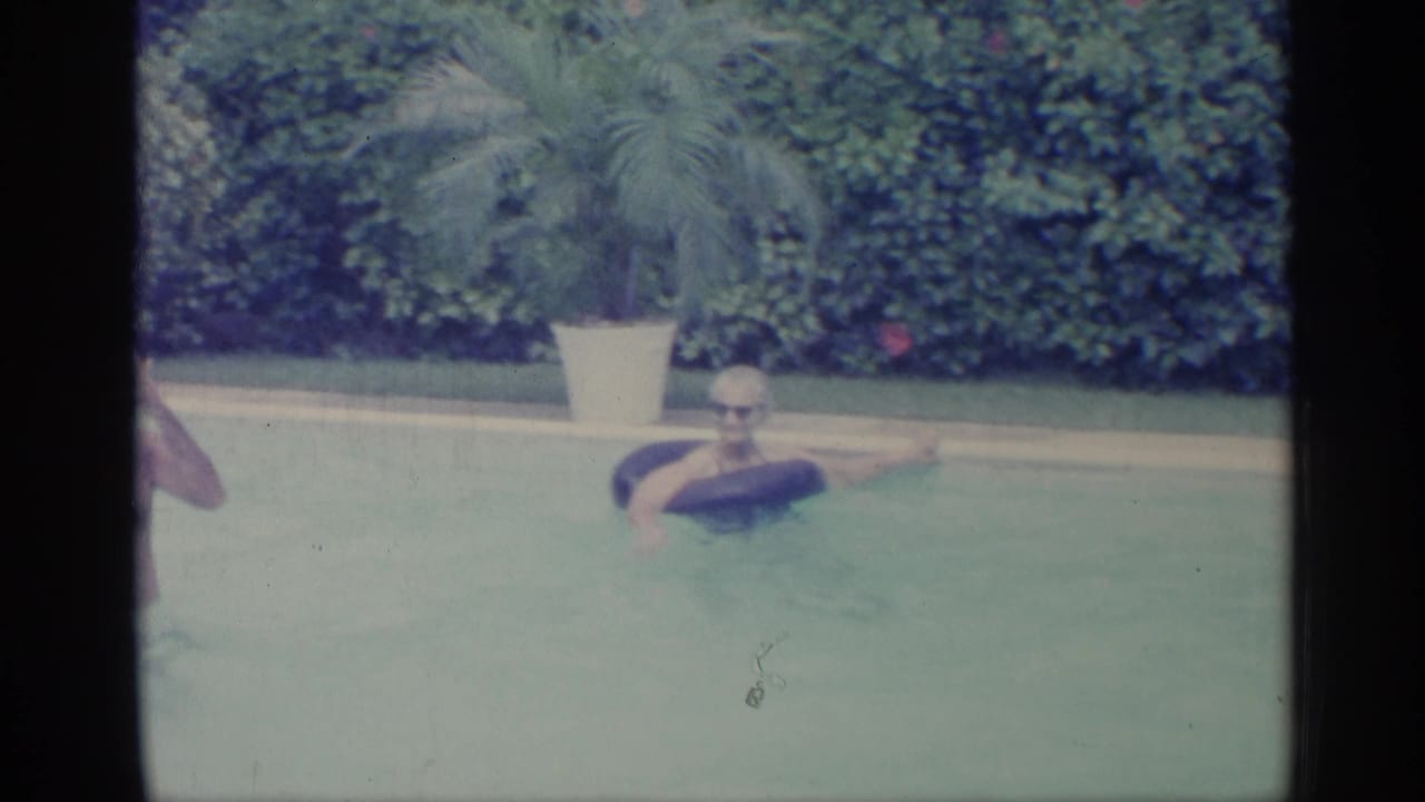1959 Indiana home-movie frame of a man on a black inner tube in a backyard pool, with another swimmer partly visible and hedges behind.