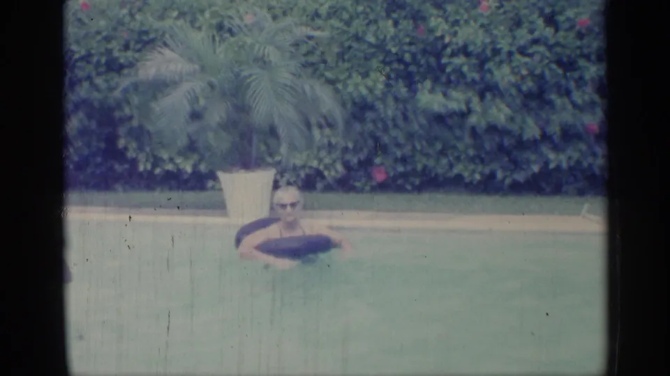 1959 Indiana 8mm still showing a man floating on a black inner tube in a backyard pool beside a potted palm and dense hedge.