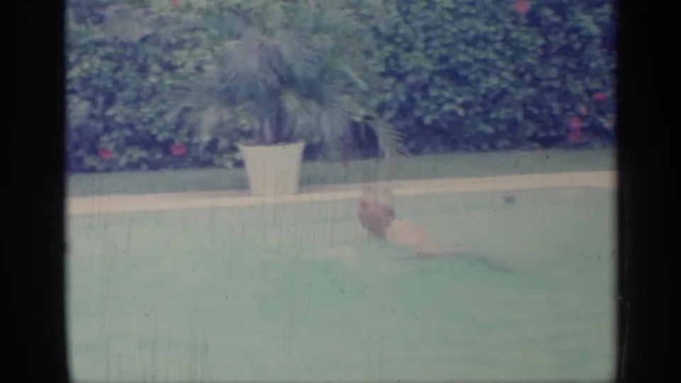 1959 Indiana home-movie frame with an adult swimmer partly submerged in a backyard pool, framed by a hedge and potted palm.
