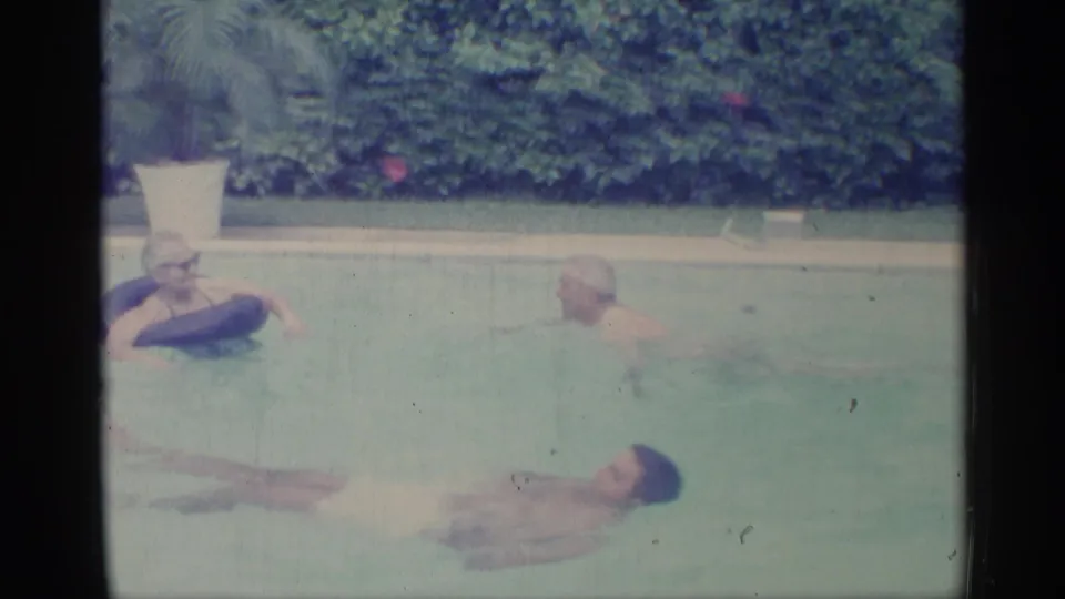 1959 Indiana home-movie frame showing adults in a pool, with one woman using a dark inflatable ring near hedges and pale pool edging.