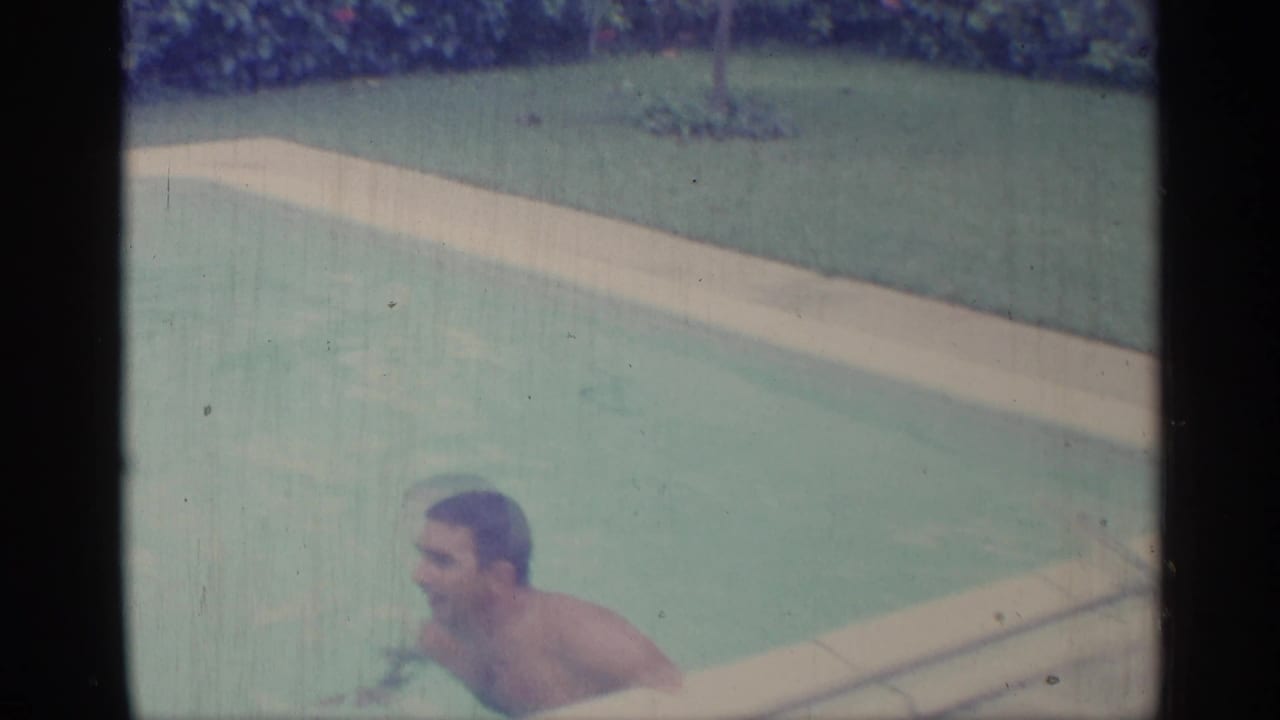 Man near the edge of a swimming pool with lawn and hedges behind him.